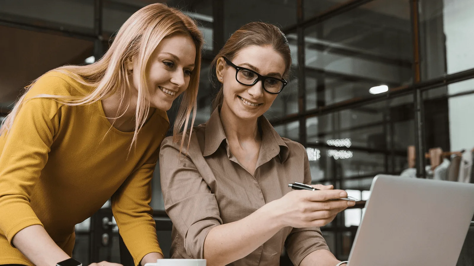 Zwei Frauen im Büro an Laptop, eine in gelbem Pullover, eine in beiger Bluse mit Brille, beide lächelnd