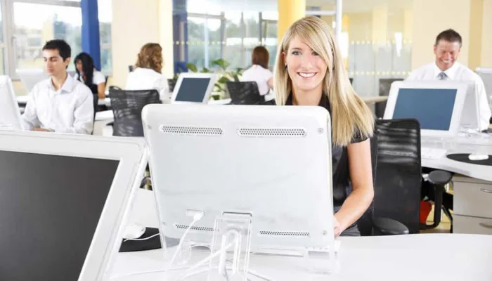 Blonde Frau am Computer im hellen Büro mit Kollegen im Hintergrund, Text 'Nordic Consulting GmbH - Läuft dein Unternehmen auc