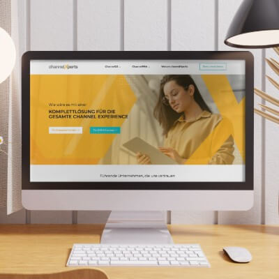 Monitor zeigt Website mit gelb-orangem Hero-Bereich und Frau mit Tablet