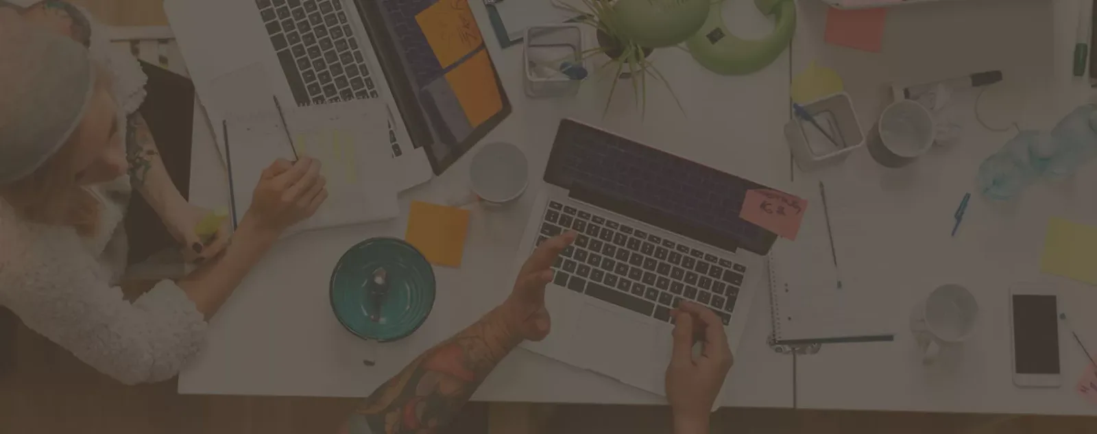 Overhead-Aufnahme eines Arbeitsplatzes mit Laptop, Händen, Pflanze und Notizbüchern