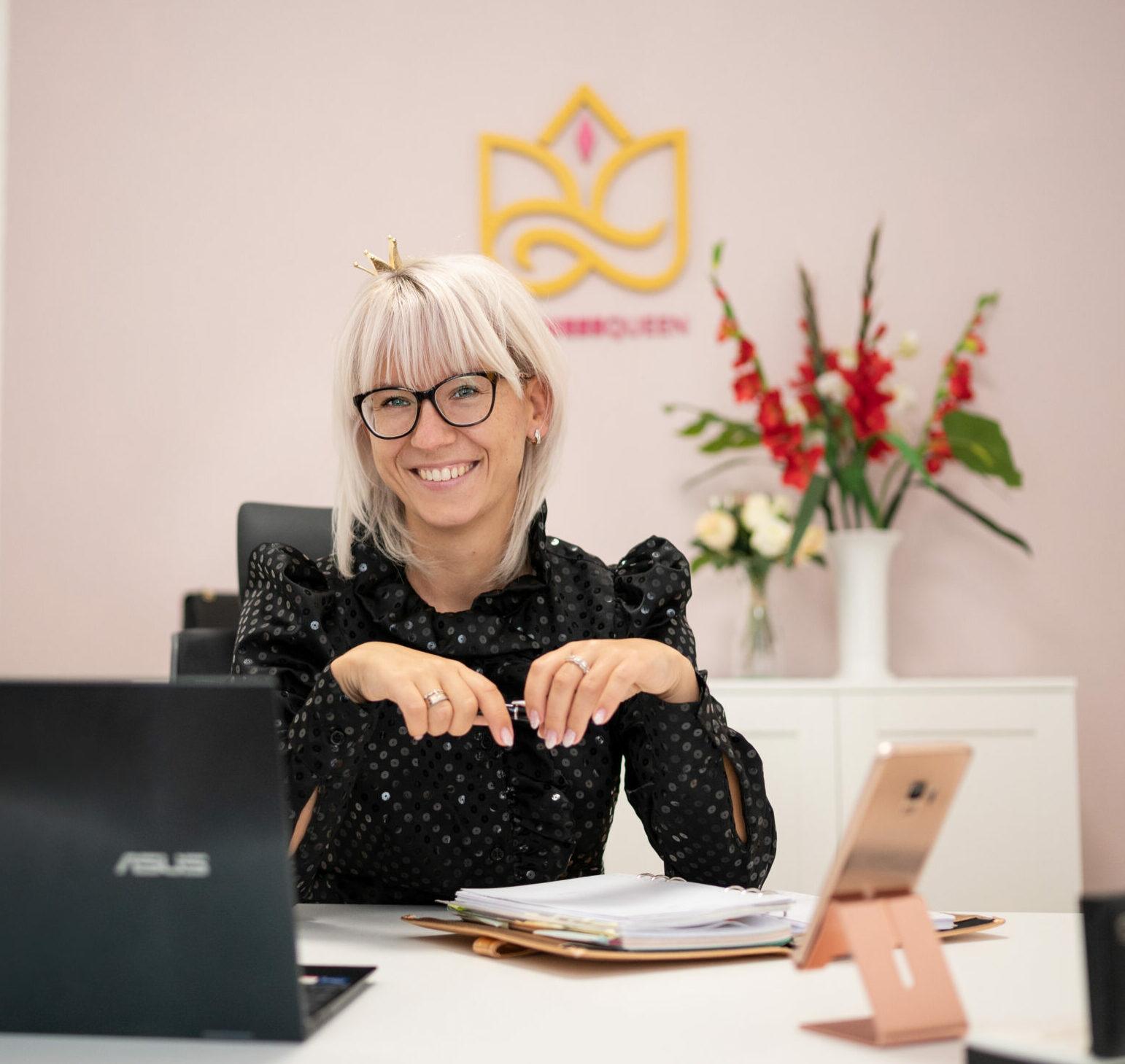 Porträt einer Frau mit weißem Haar und Brille am Schreibtisch vor gelbem Logo