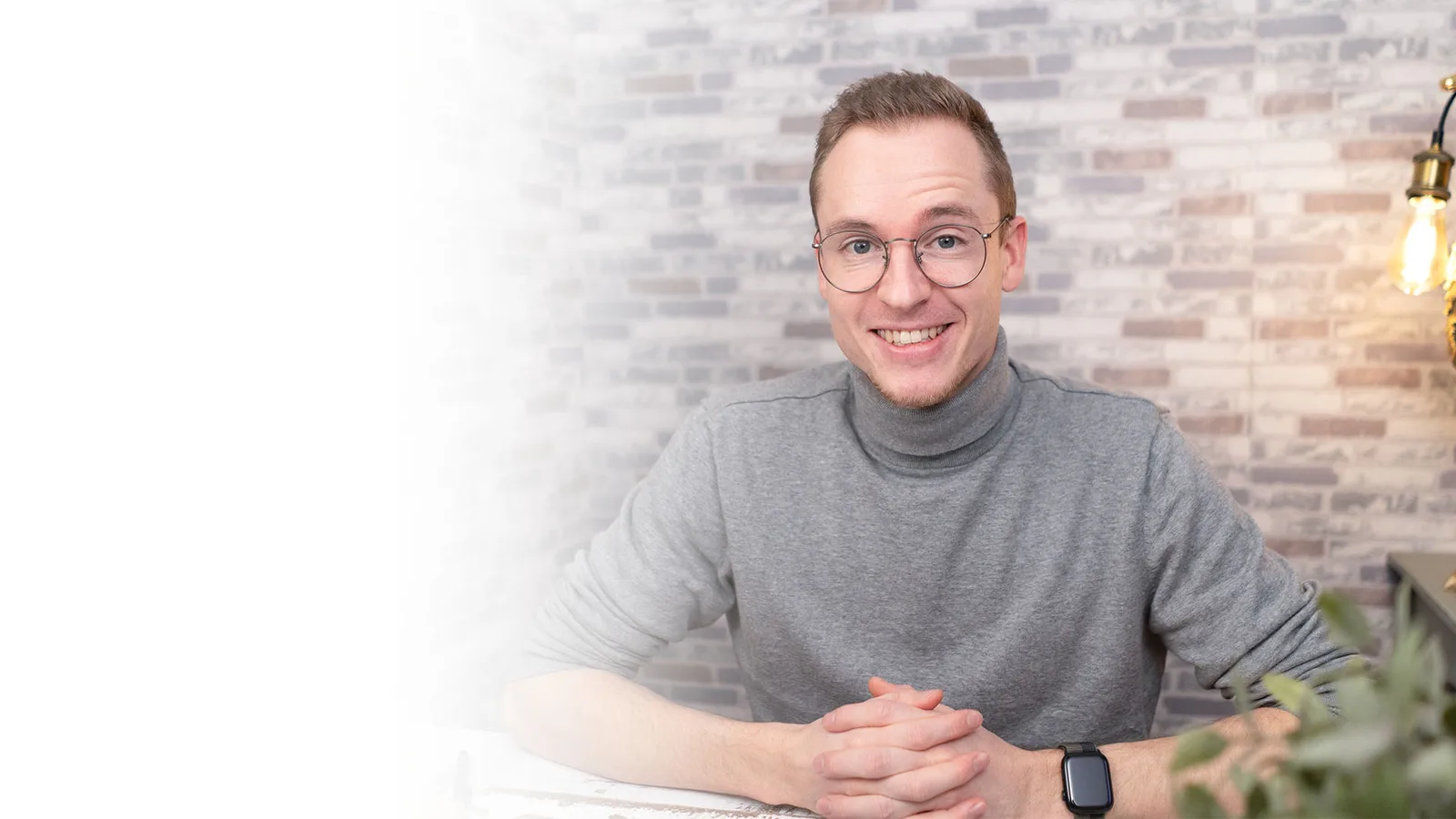 Timo Ostrich in grauem Rollkragenpullover vor Backsteinwand, lächelnd am Schreibtisch sitzend
