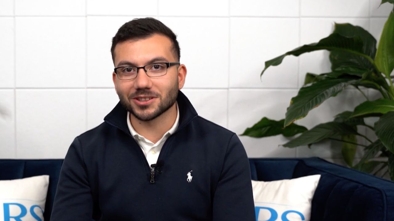 Mann mit Brille sitzt auf Sofa vor weißer Wand, lächelt in die Kamera