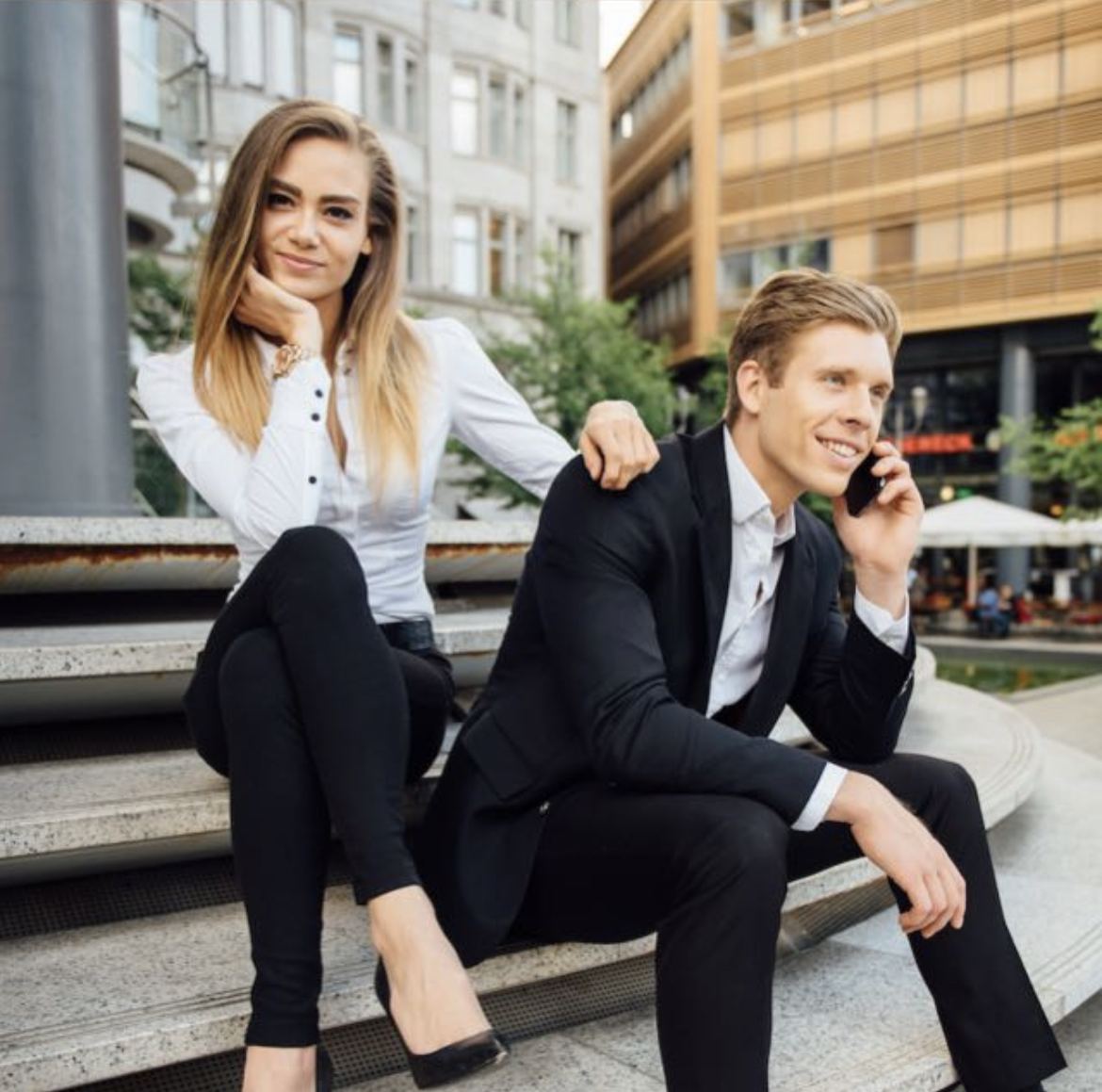 Sabrina Nennemann und Fabian Durek sitzen auf Stufen in einer urbanen Umgebung, beide in Business-Kleidung