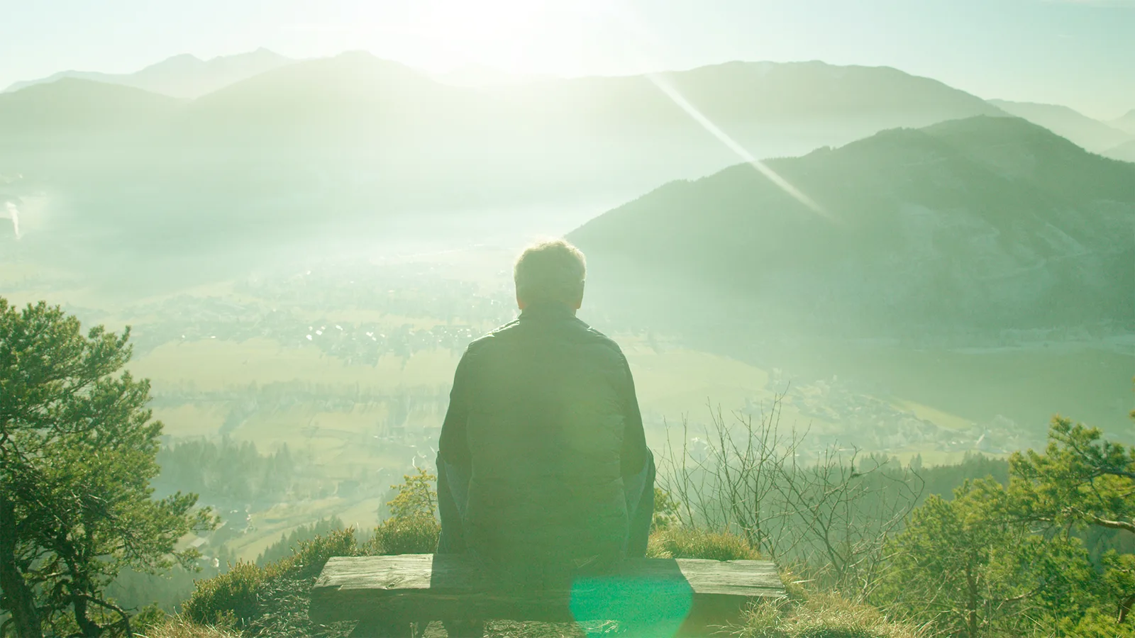 Person von hinten auf Bank mit Ausblick auf Berge und Tal, grüne Farbstimmung, inspirierende Landschaft