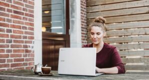 Frau mit Laptop auf Holztisch vor Ziegelmauer, Kaffee, nachdenklich