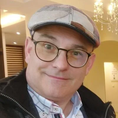 Portrait eines Mannes mit Brille und Kappe, lächelnd, vor indoorlem gelbem Hintergrund