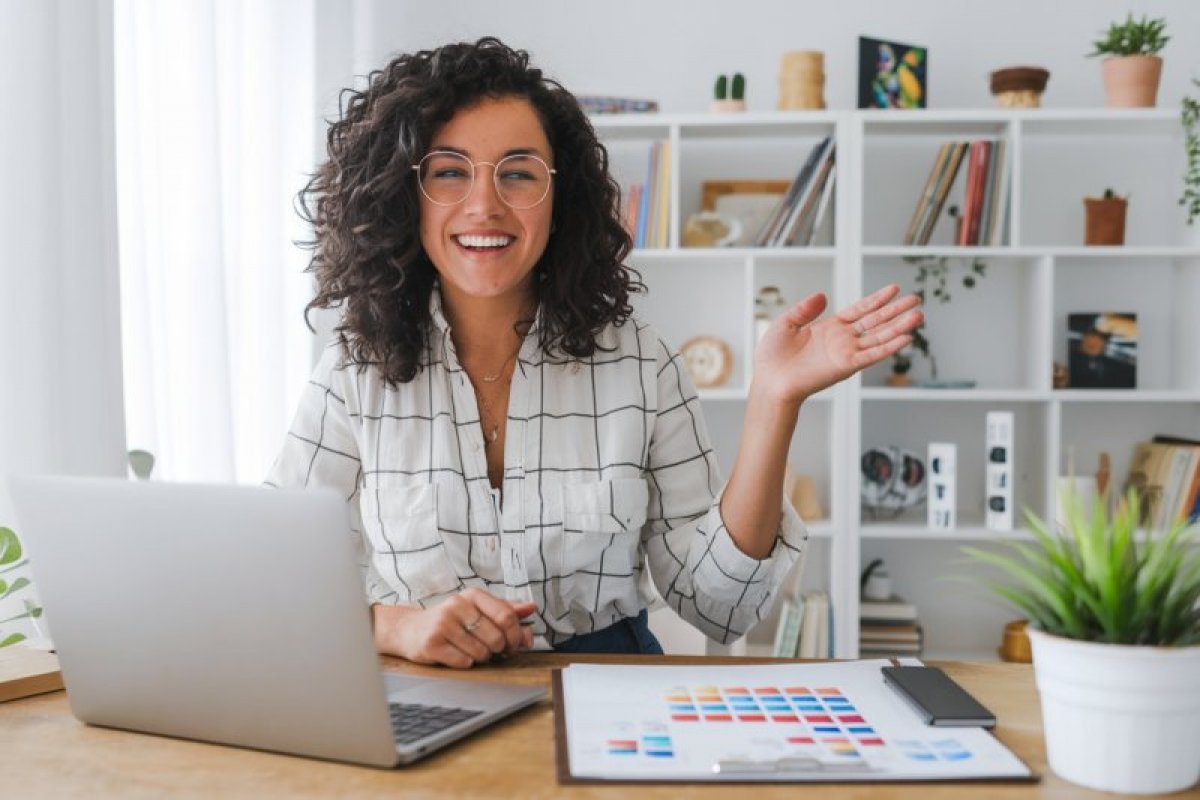 Junge Frau mit Brille am Laptop lächelnd, Bücherregal im Hintergrund, positive Ausstrahlung