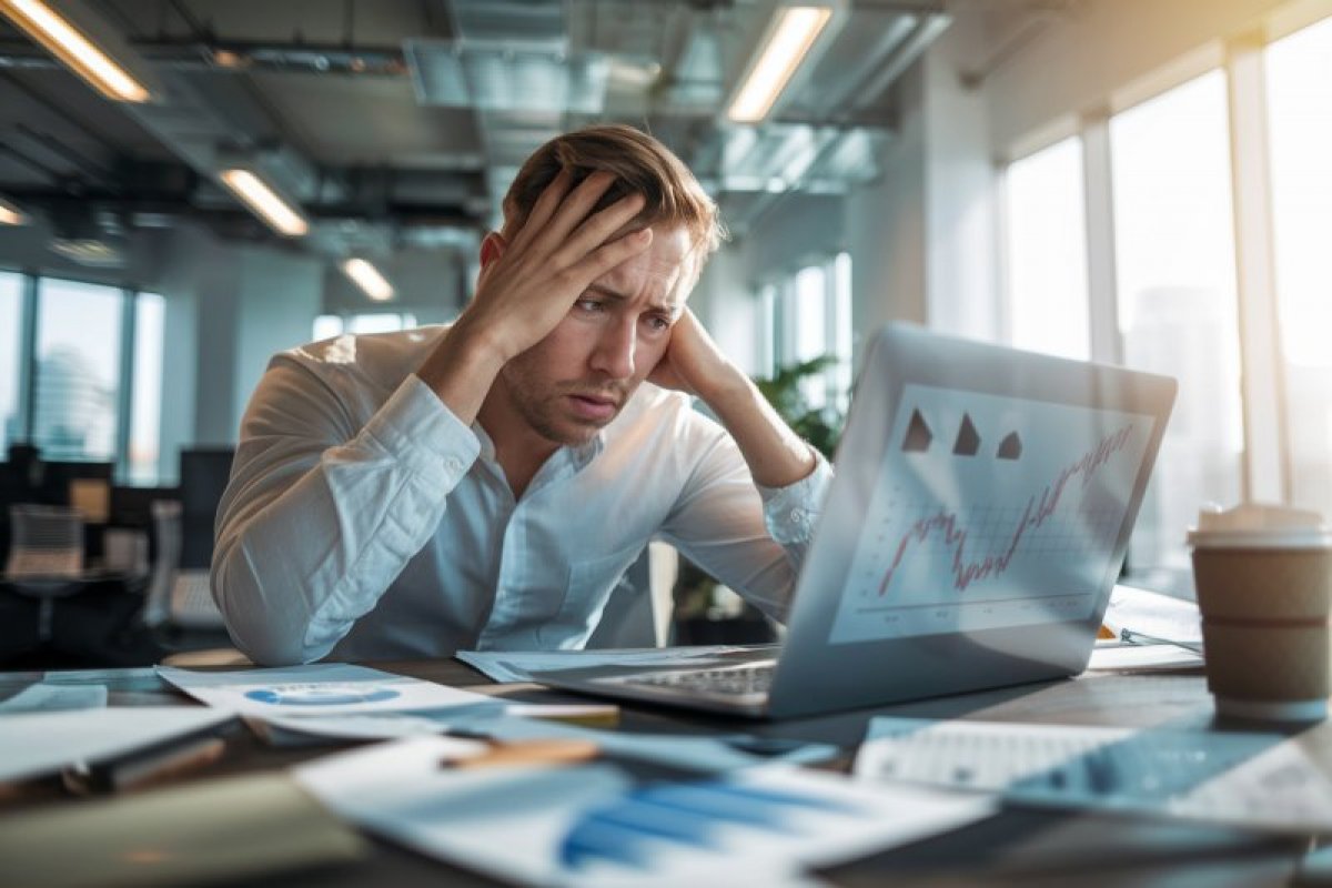 Gestresster Mann am Laptop mit Datenblättern auf dem Schreibtisch, nachdenkliche Pose