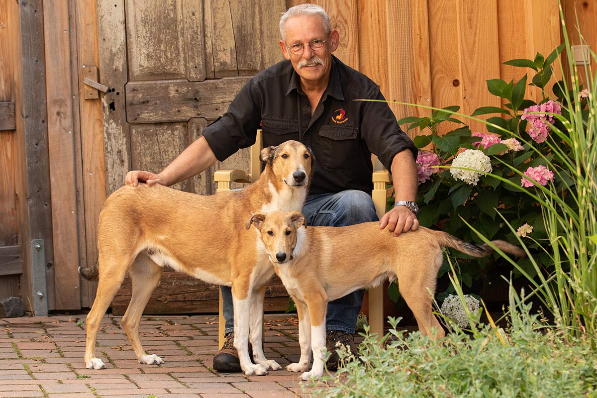 Jan Nijboer sitzt mit zwei Pharaoh Hound oder ähnlichen Hunderassen vor Holztür