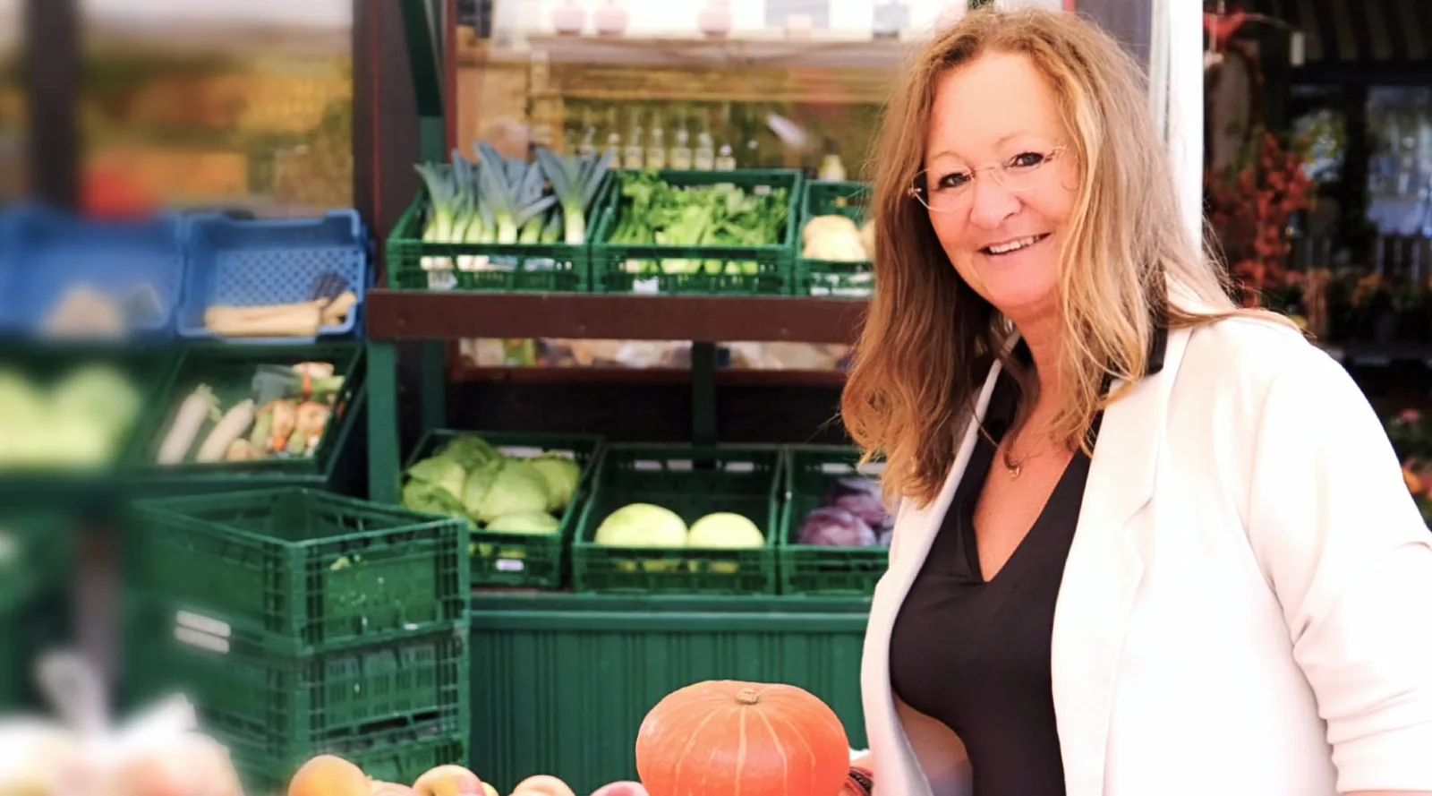 Birgit Lieser lächelt am Gemüsestand mit grünen Boxen voller frischem Gemüse und Kürbis im Vordergrund