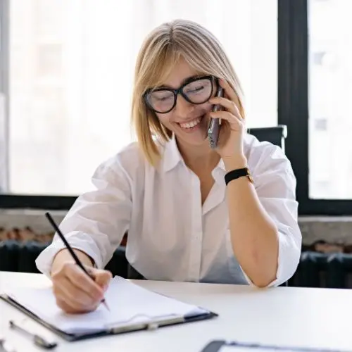 Das perfekte Verkaufsgespräch – Vertriebstraining als Videokurs 4 Porträt: Junge Frau in weißem Hemd mit Brille am Telefon, schreibend am Schreibtisch