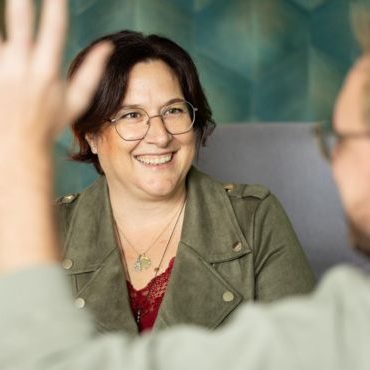 Marlen Kästner in olivfarbener Jacke bei Gruppengespräch, lächelnd mit Brille