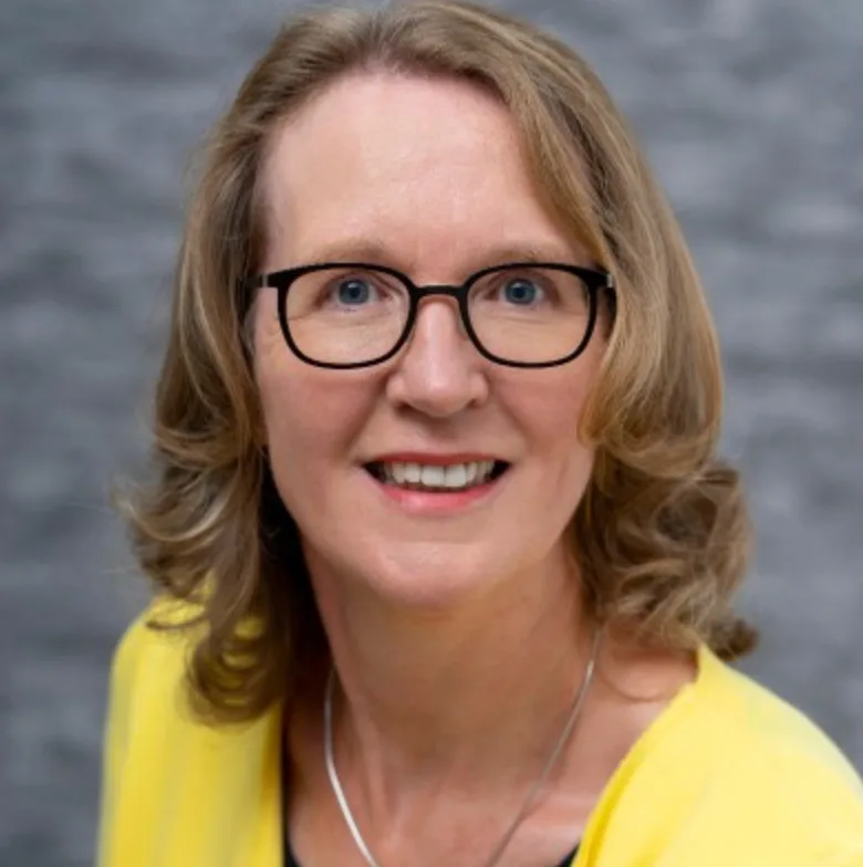 Frau mit schulterlangen hellbraunen Haaren, schwarzer Brille und gelbem Oberteil lächelnd