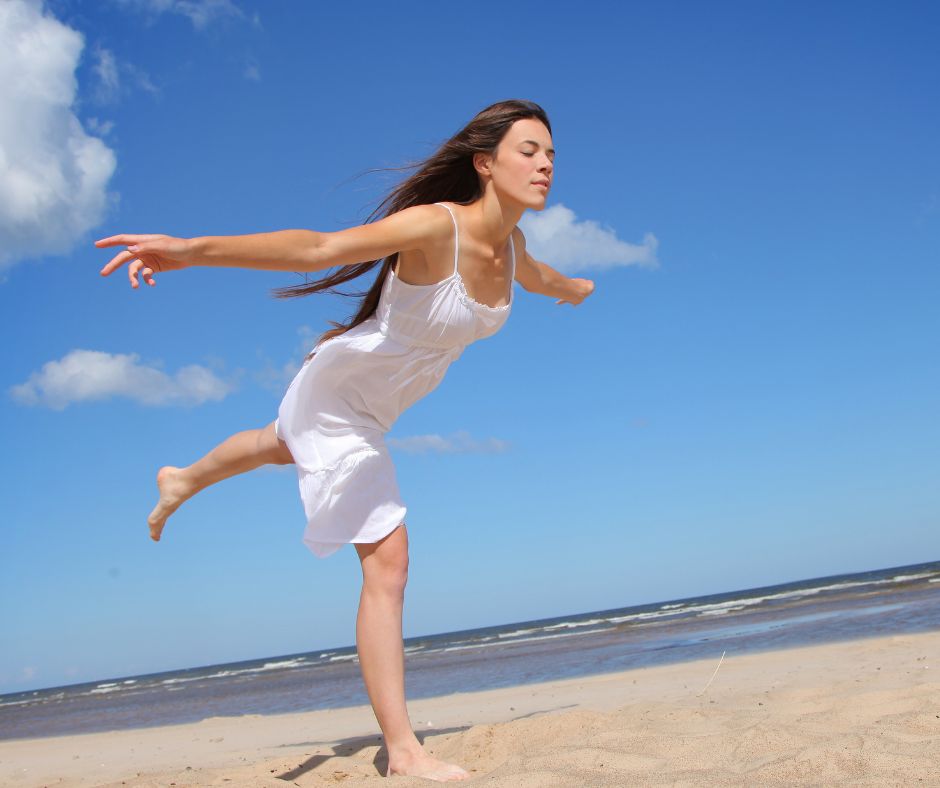 Frau in weißem Kleid springt am Strand vor blauem Himmel, Arme ausgebreitet