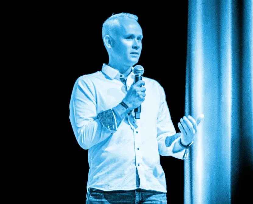 Andreas am Rednerpult mit Mikrofon vor schwarzem Hintergrund, blaue Ausleuchtung