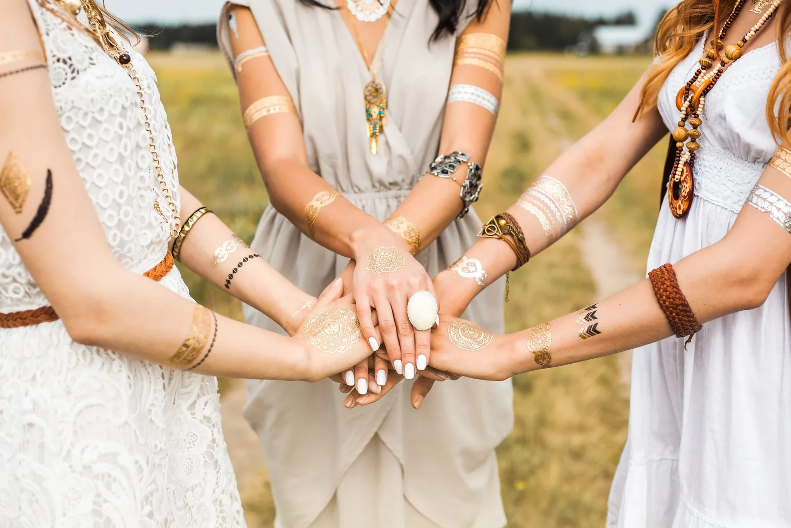 Drei Frauen zeigen ihre Hände mit Boho-Flash-Tattoos, Ringen und Armbändern auf grüner Wiese