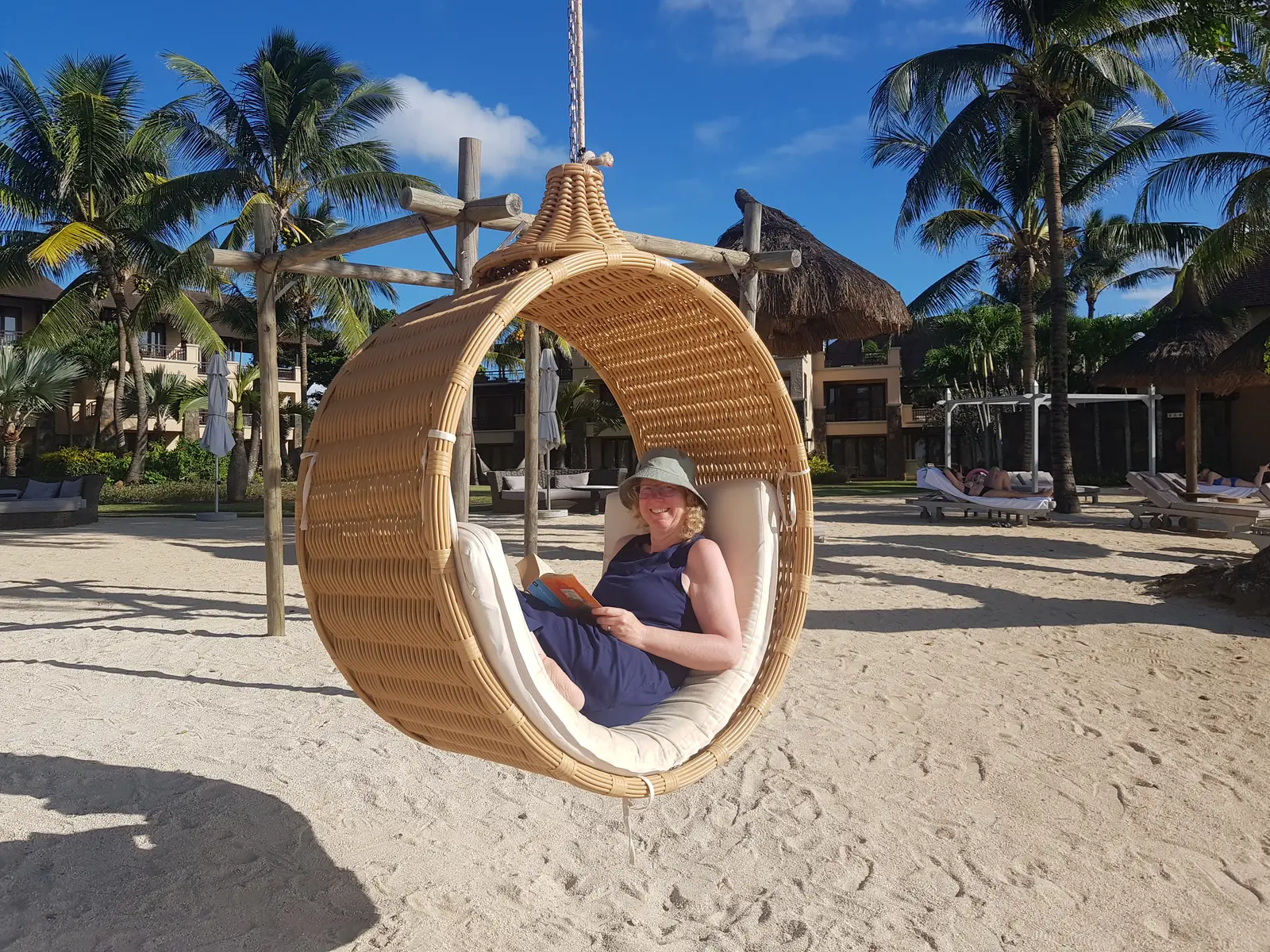 Frau sitzt entspannt in hängender Korbschaukel am Strand mit Palmen und blauem Himmel, liest Buch