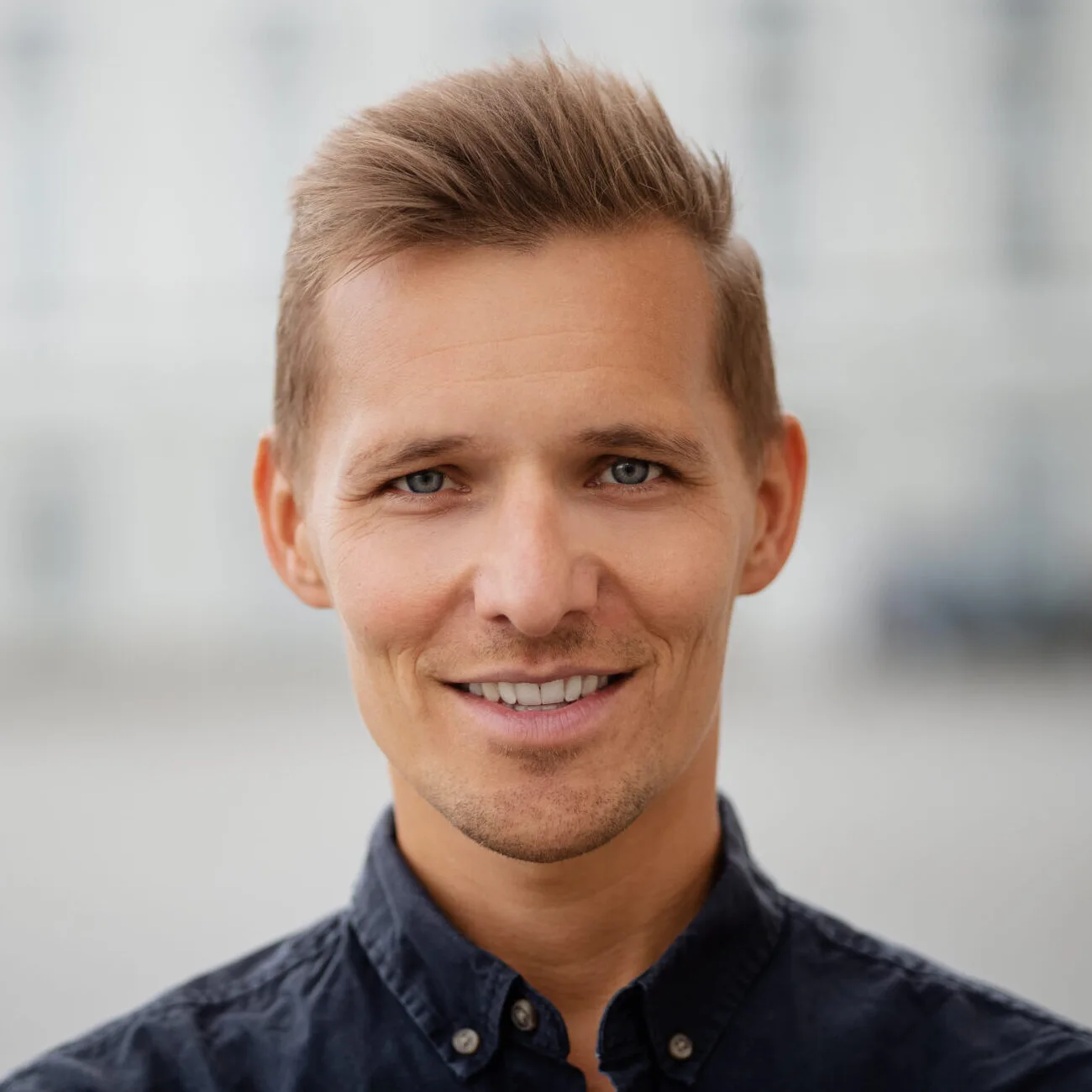 Junger Mann mit hellblondem Haar und Schnurrbart, schwarzem Oberteil, lächelnd vor unscharfem Hintergrund