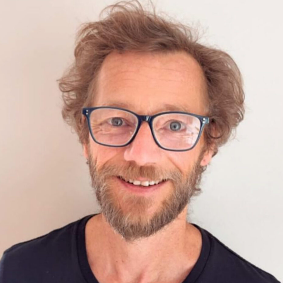 Mann mit lockigem braunem Haar, Brille und braunem Bart, schwarzem Oberteil vor hellem Hintergrund