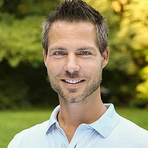 Mann mit dunklem Haar, hellblauem Hemd, lächelnd vor grünem unscharfem Hintergrund