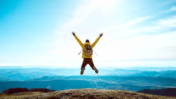 Person in gelbem Rucksack springt jubelnd auf Berg mit Blick auf Landschaft