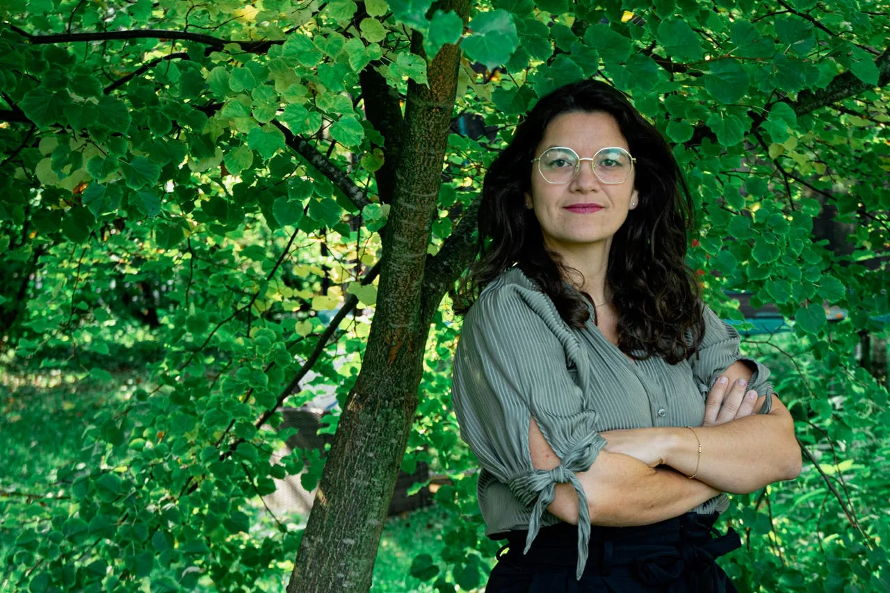 Der Gartenkongress – Gärtnern und Ernten im Herbst 7 Frau mit schwarzem Haar, Brille und grauem Hemd, verschränkte Arme vor grünem Laub und Baumstamm