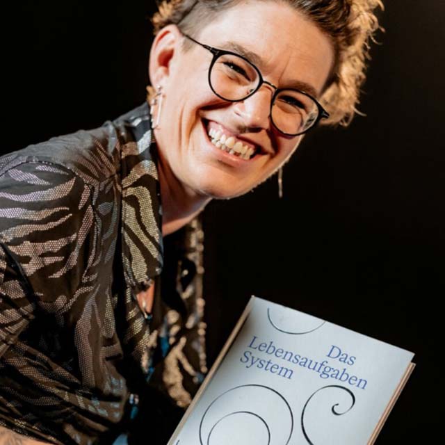 Frau mit Brille und Zebra-Muster-Jacke hält Buch Lebensaufgaben System und lächelt