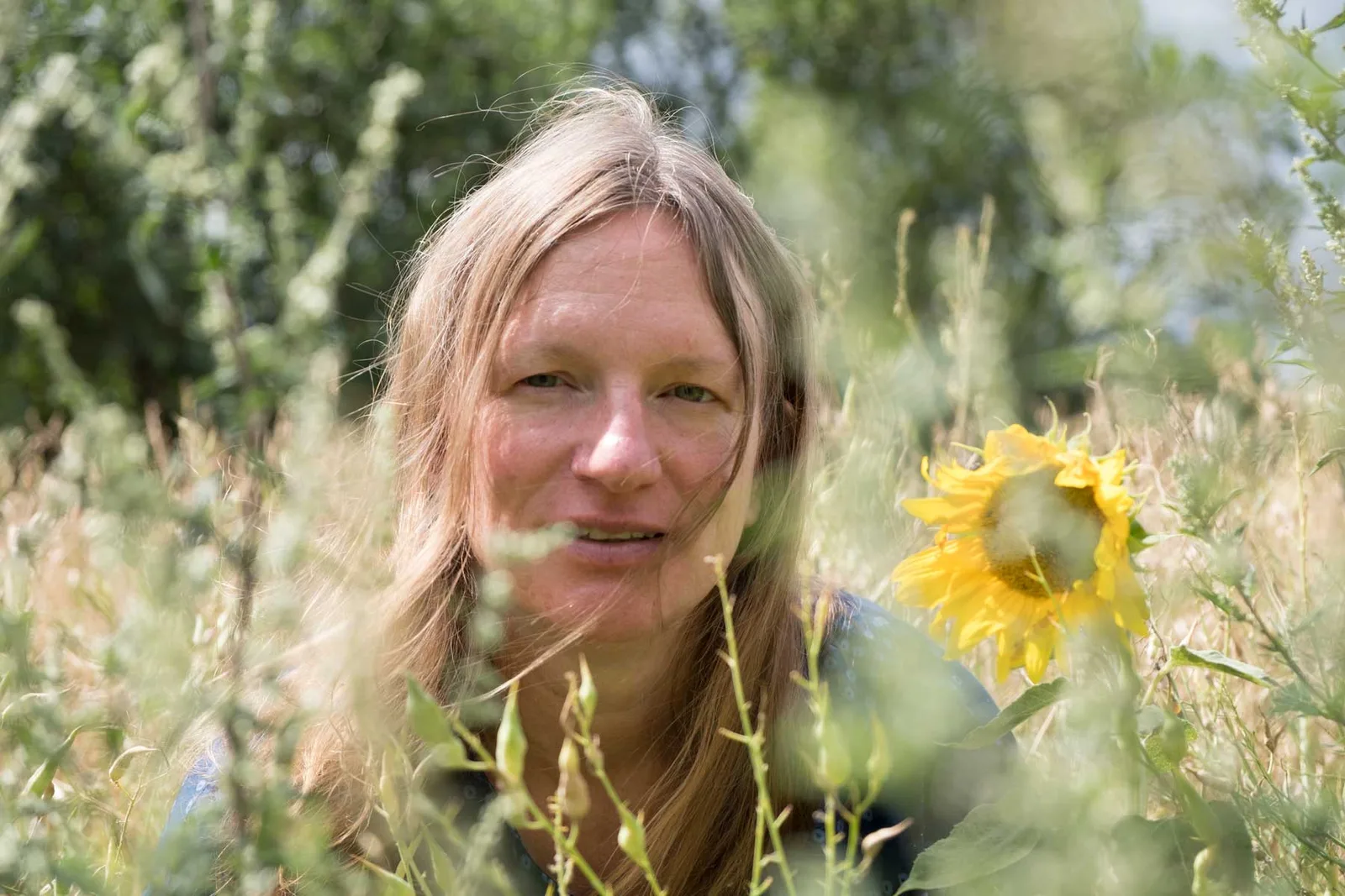 Silke Galla lächelt inmitten von grünen Pflanzen und gelben Blüten im Garten