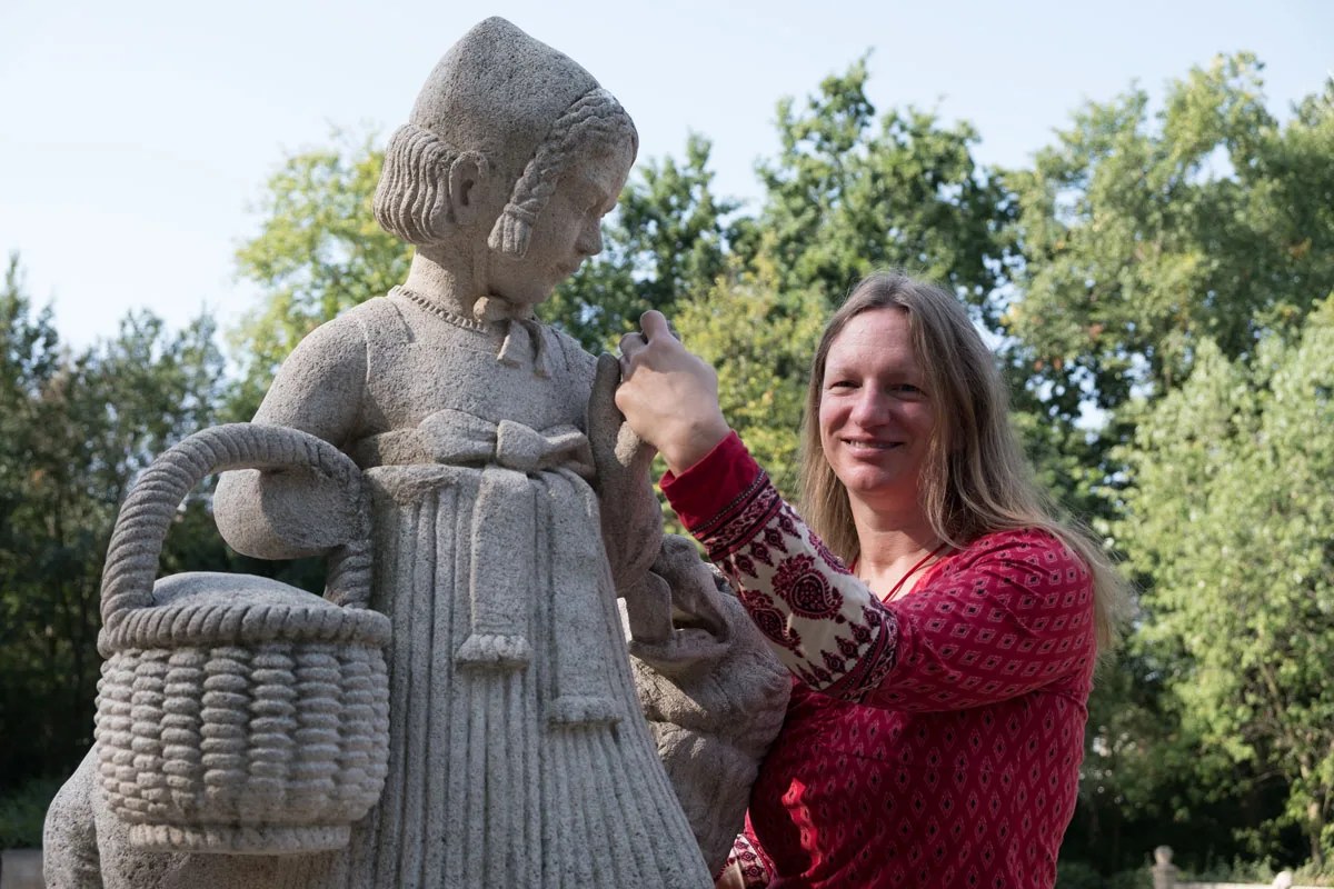 Silke Galla posiert lächelnd neben einer Steinstatue einer Frau mit Korb