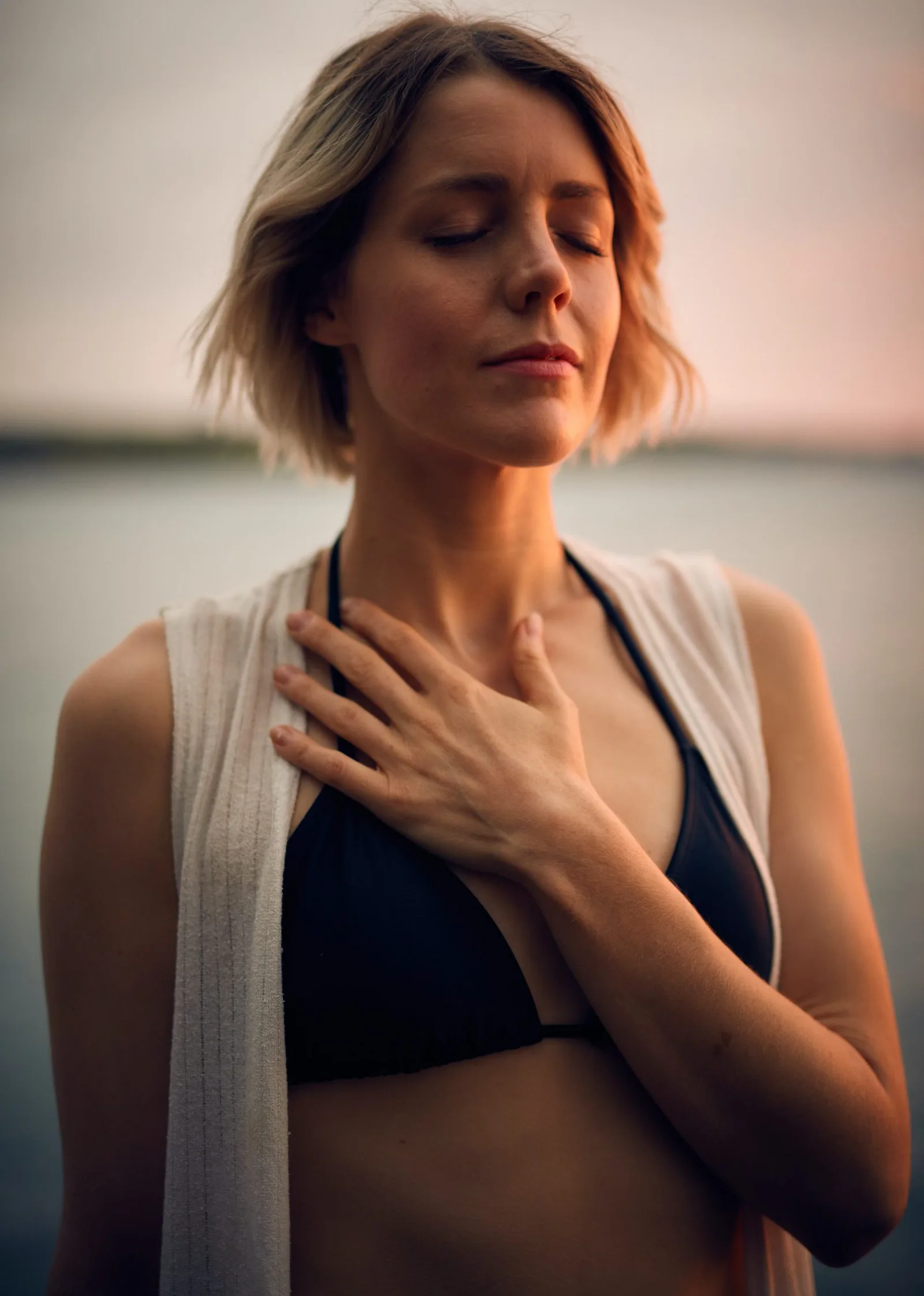 Frau in schwarzem Bikini und weißem Cardigan mit Hand auf Brustbereich, Seeufer im Hintergrund