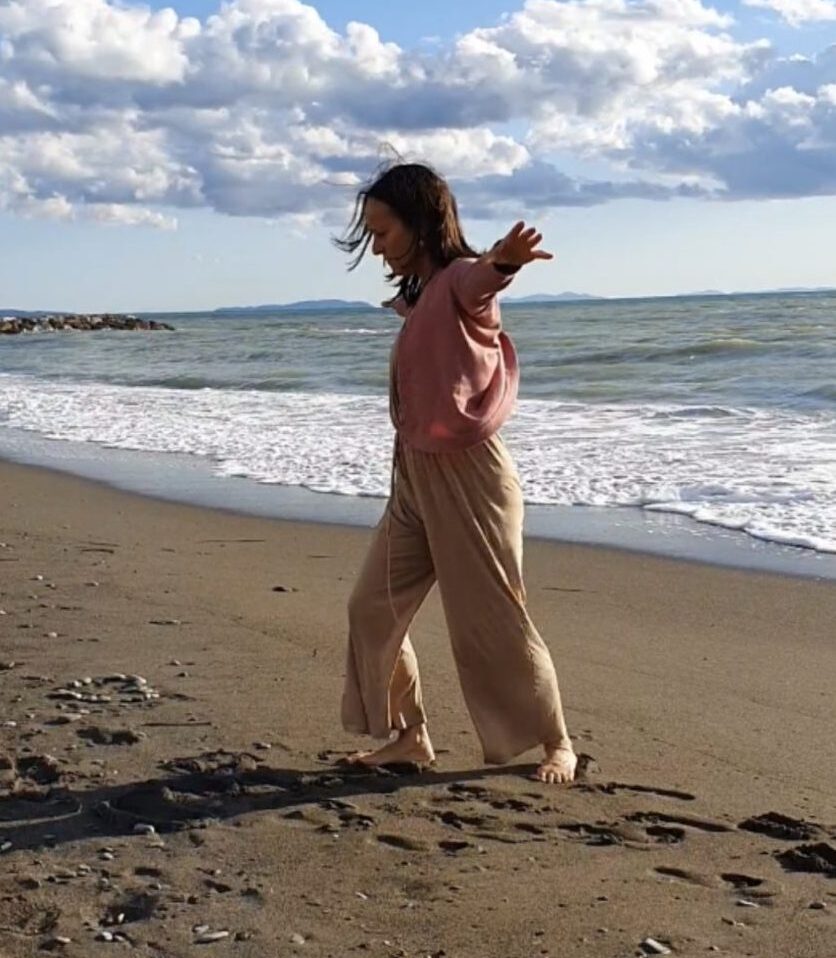 Frau in roséfarbenem Oberteil und beigefarbener Hose mit ausgestrecktem Arm am Strand