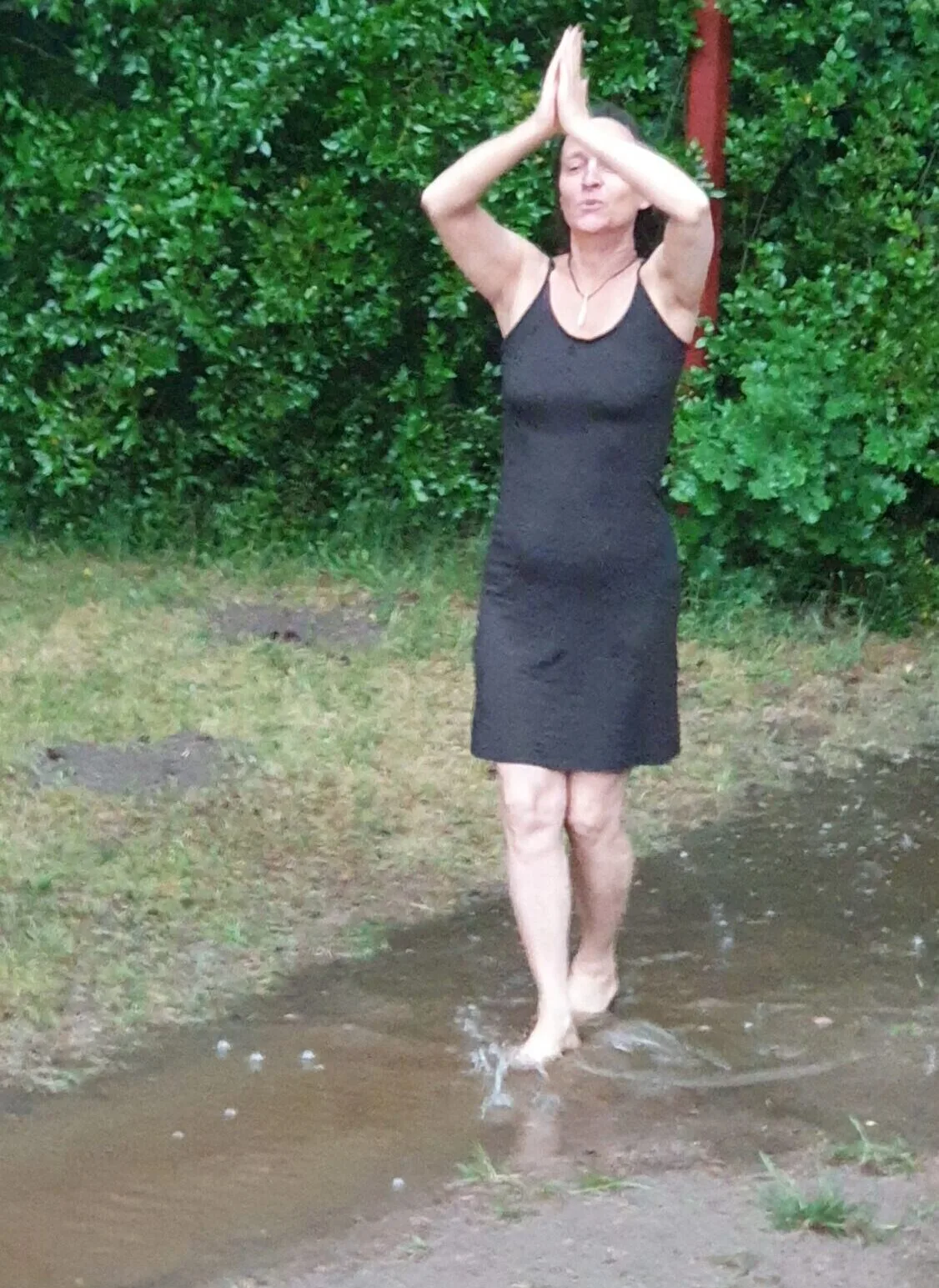 Frau in schwarzem Kleid springt in Pfütze vor grüner Hecke