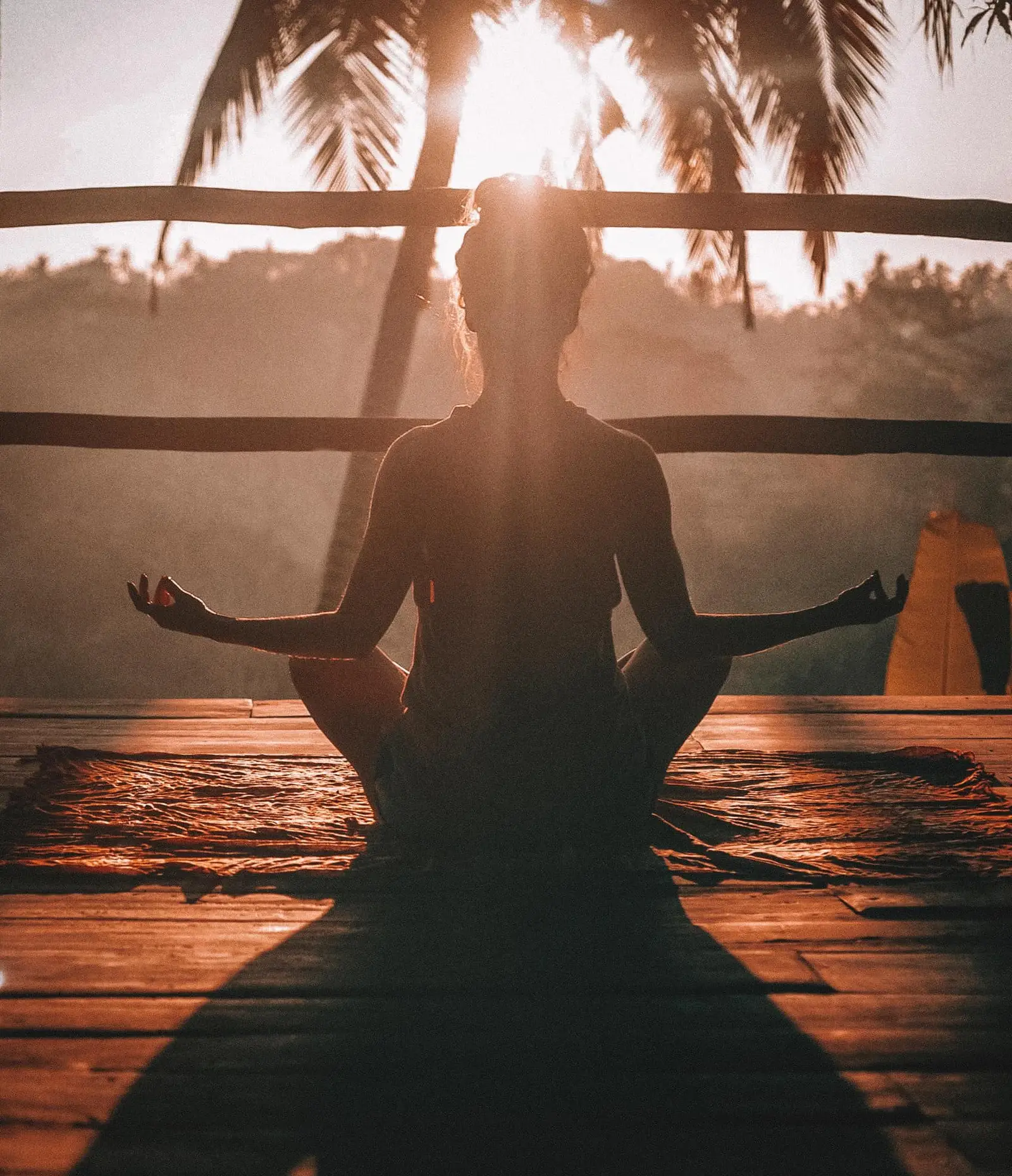 Die AcroYoga Uni – Mit Spiel & Spaß AcroYoga lernen 21 Silhouette einer Person in Meditationshaltung auf Holztegen bei Sonnenuntergang mit Palmenbäumen und Meer im Hintergrund
