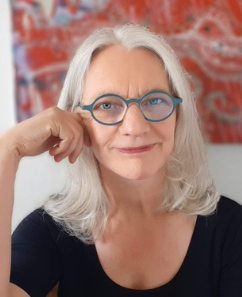 Andrea Brummack, Frau mittleren Alters mit weißem Haar und Brille, vor rötlichem Hintergrund