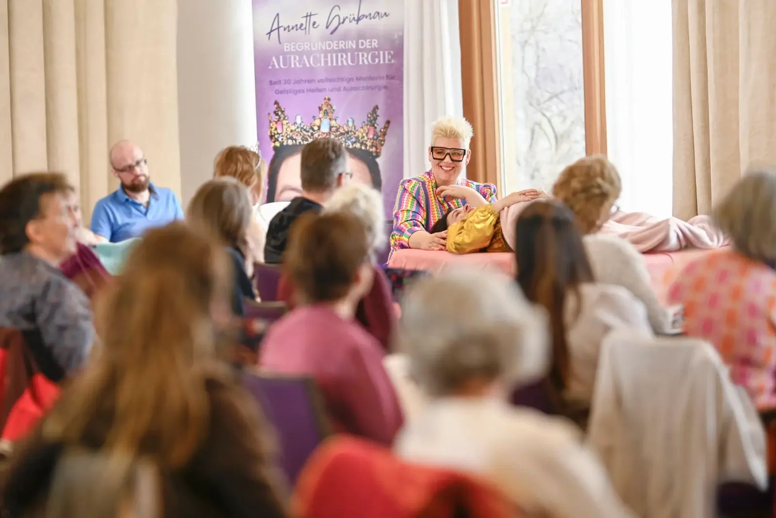 Annette Grübnau conducting a seminar in colorful jacket and white-framed glasses, presenting to seated participants, with lar