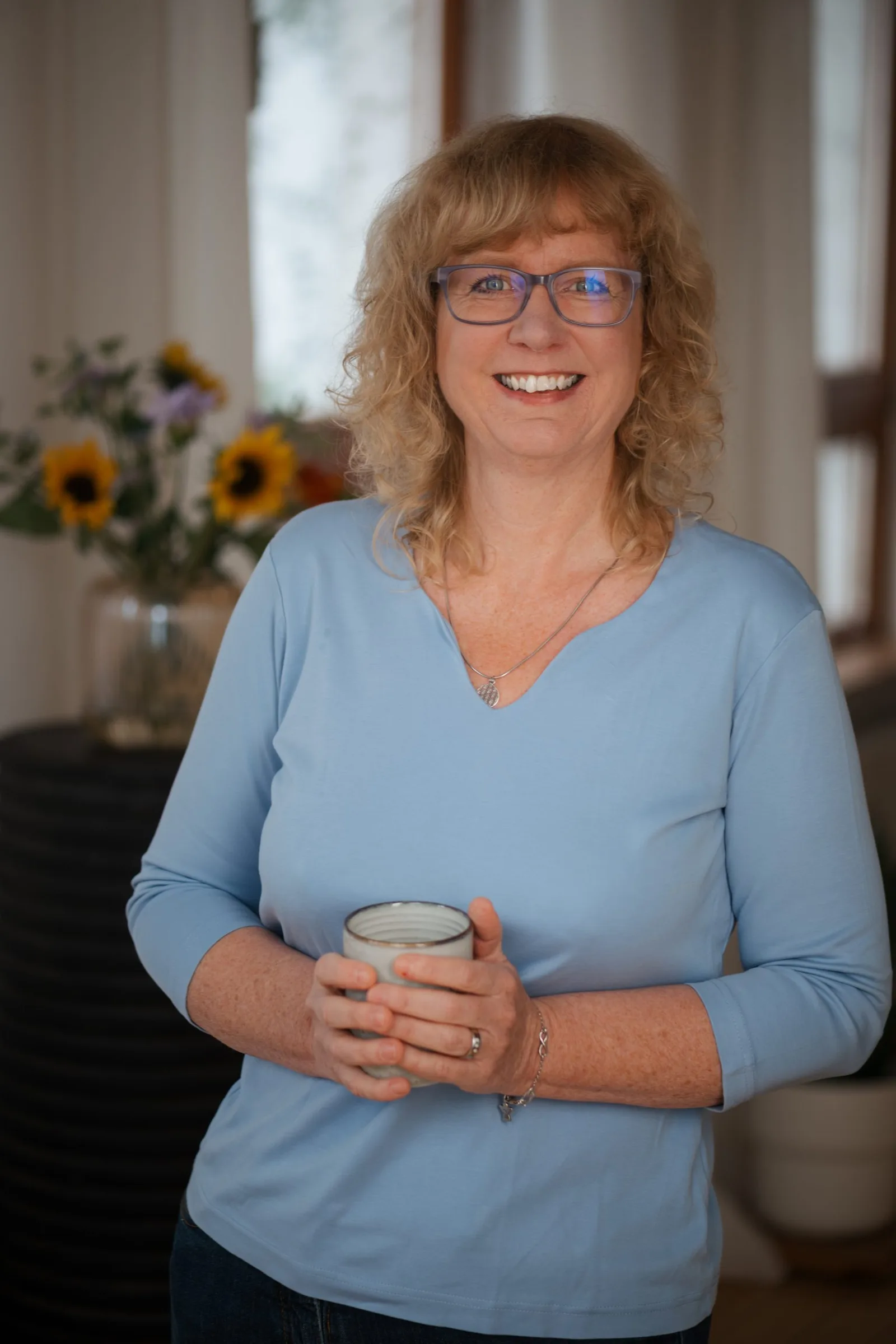 Porträt einer Frau mit welligem rötlichblondem Haar, Brille, helles blaues Langarmshirt, hält Kaffeetasse, freundliches Läche