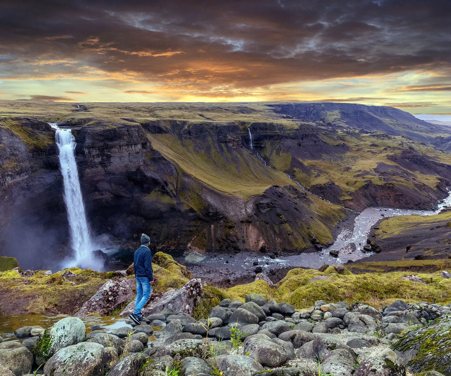 Person in blauer Jacke schaut auf Wasserfall und isländische Landschaft