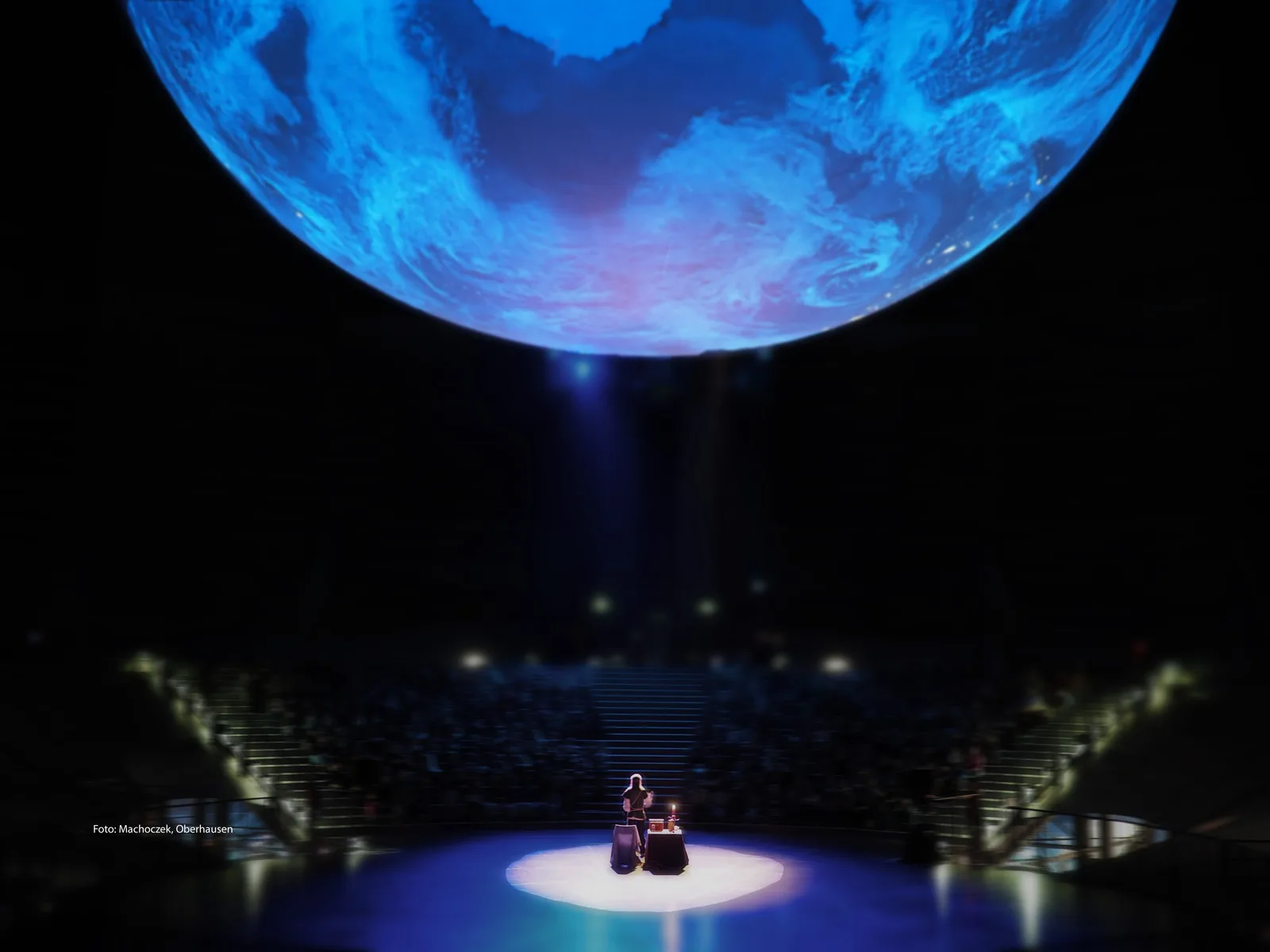 Theaterbühne mit blauem Planeten und sitzendem Performer im Scheinwerferlicht