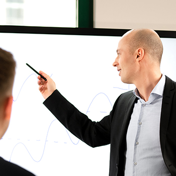 Die perfekte Altersvorsorgeberatung – Videokurs für Finanzberater 6 Robert Peukert bei Seminar, zeigt auf Flipchart mit Grafik, dunkler Anzug, hellblaues Hemd