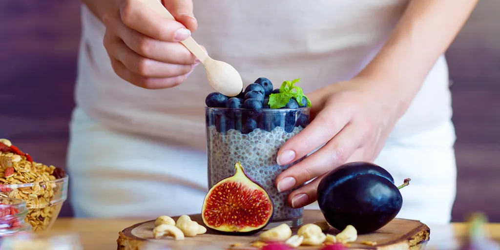 Frauenhände halten ein Glas mit Chiasamen-Pudding, Blaubeeren und Minze, mit frischen Lebensmitteln auf Holztisch