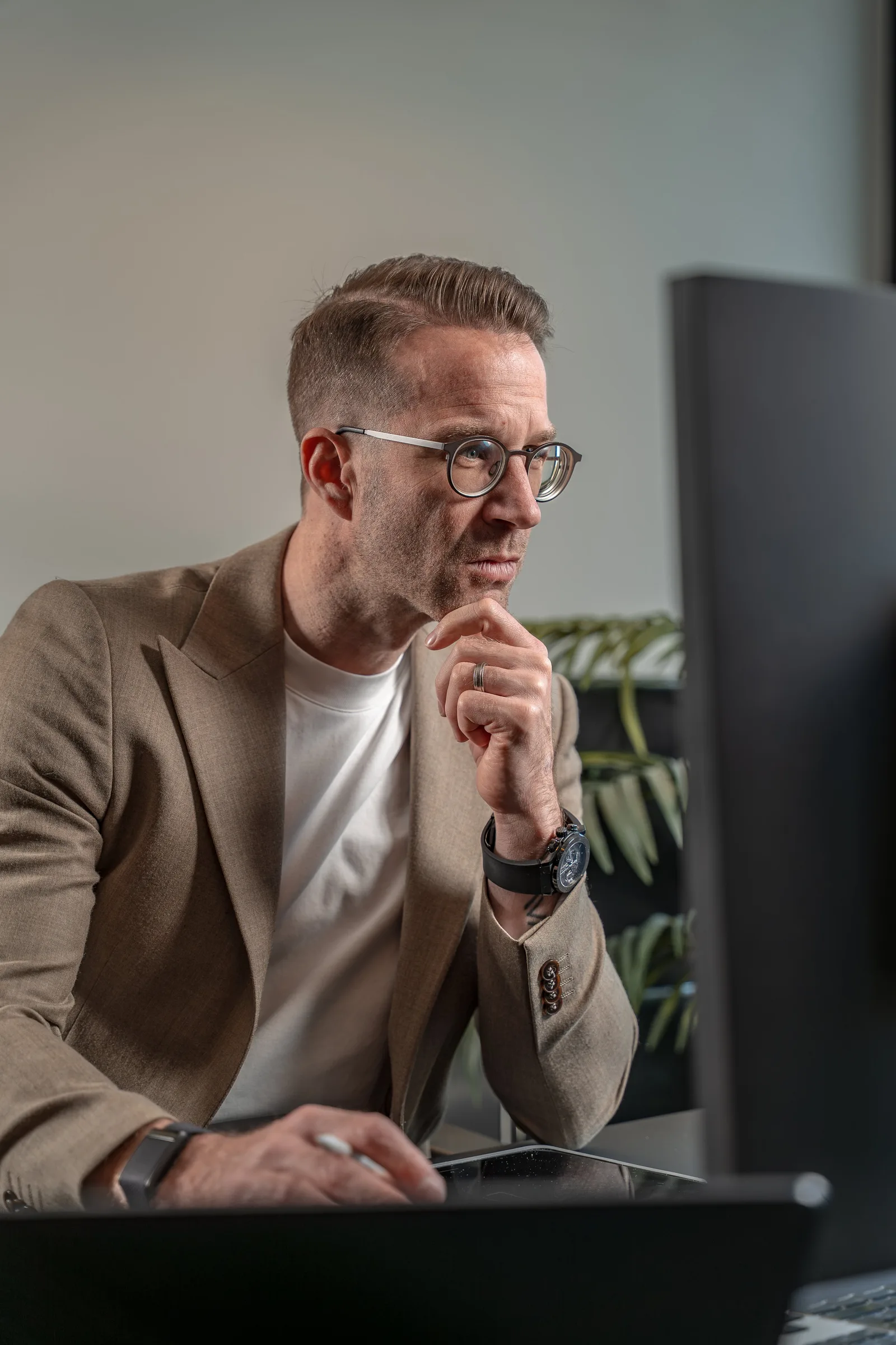 Mann mit Brille in beigem Blazer sitzt nachdenklich am Schreibtisch vor Monitor