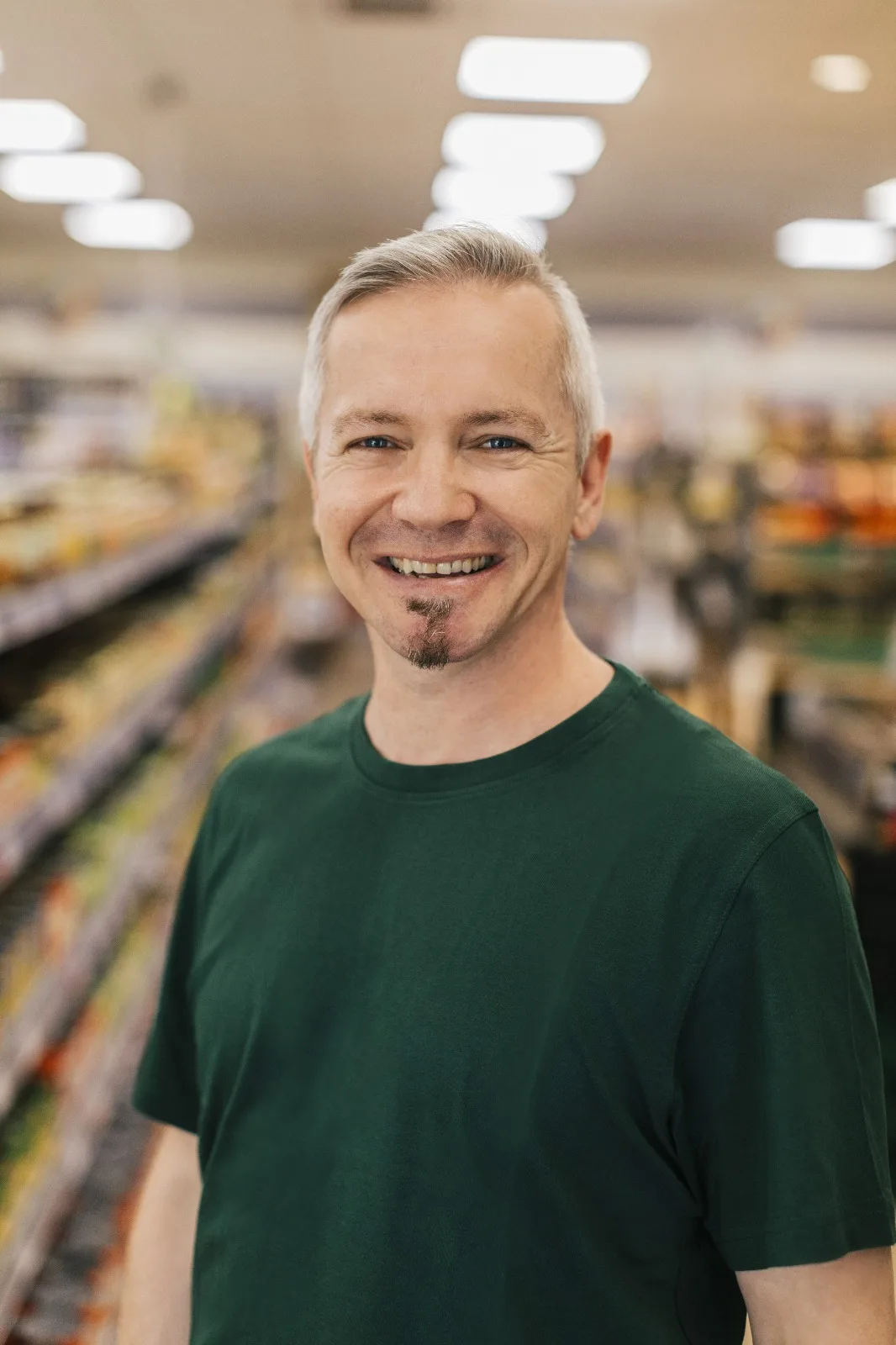 Portrait: Tim Bach, Mann mit grauem Haar und Bart in grünem T-Shirt, Supermarkt-Hintergrund