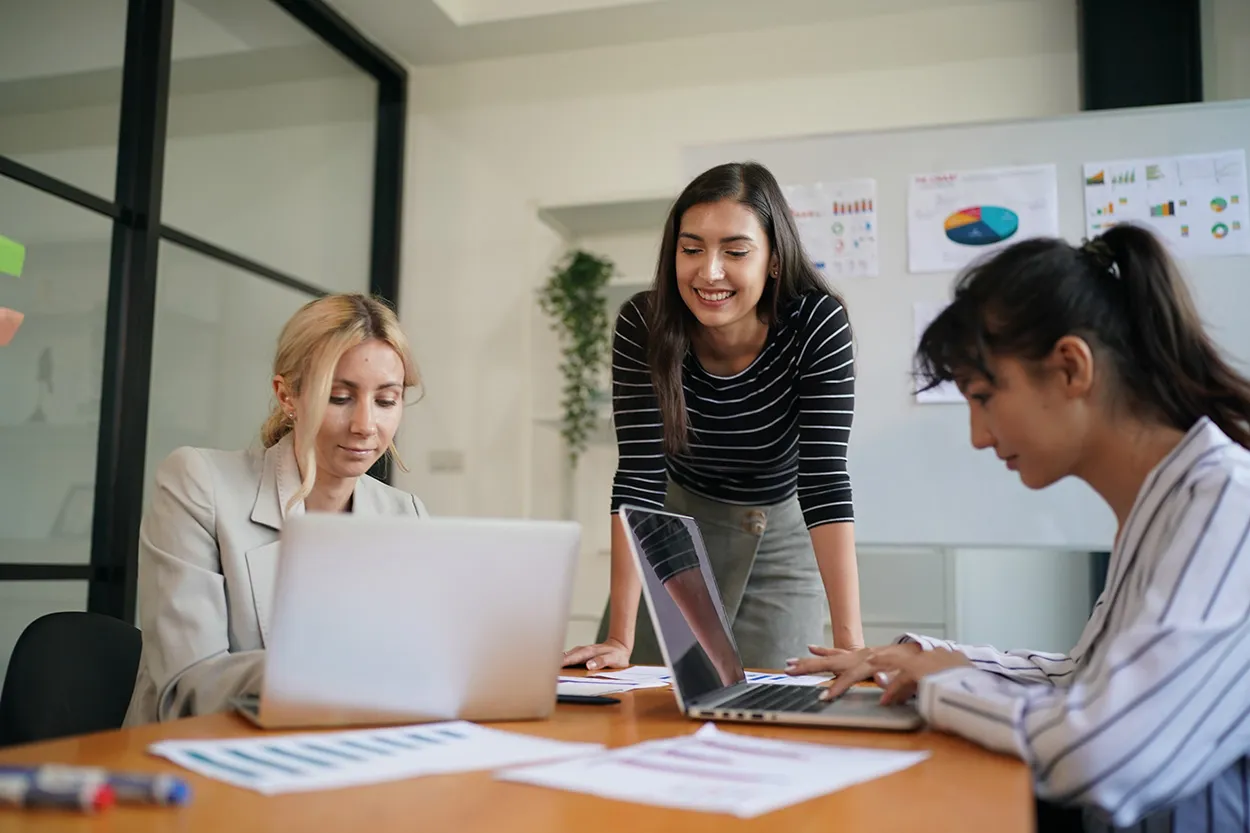 Drei Frauen arbeiten gemeinsam an Laptops in einem modernen Büro mit Whiteboard und Diagrammen