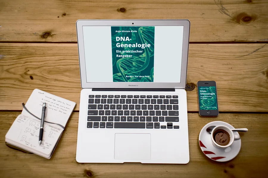 MacBook Air zeigt DNA-Genealogie Buchcover, umgeben von Notizbuch, Stift, Smartphone und Kaffeetasse