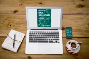 MacBook Air mit DNA-Genealogie Buchcover auf dem Bildschirm, daneben Notizbuch, Stift, Smartphone und Kaffeetasse auf Holztis
