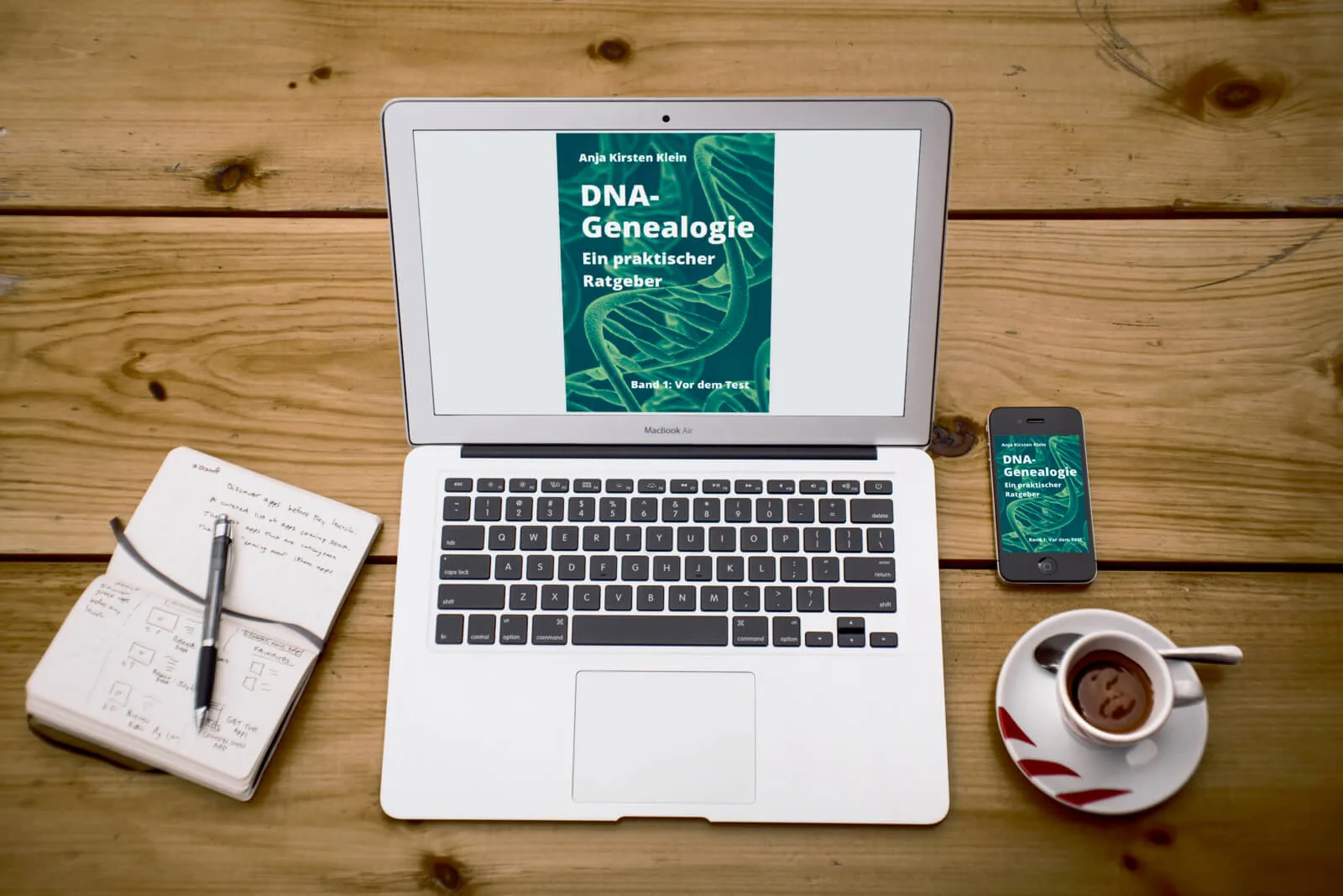 MacBook Air mit DNA-Genealogie Buchcover auf dem Bildschirm, daneben Notizbuch, Stift, Smartphone und Kaffeetasse auf Holztis
