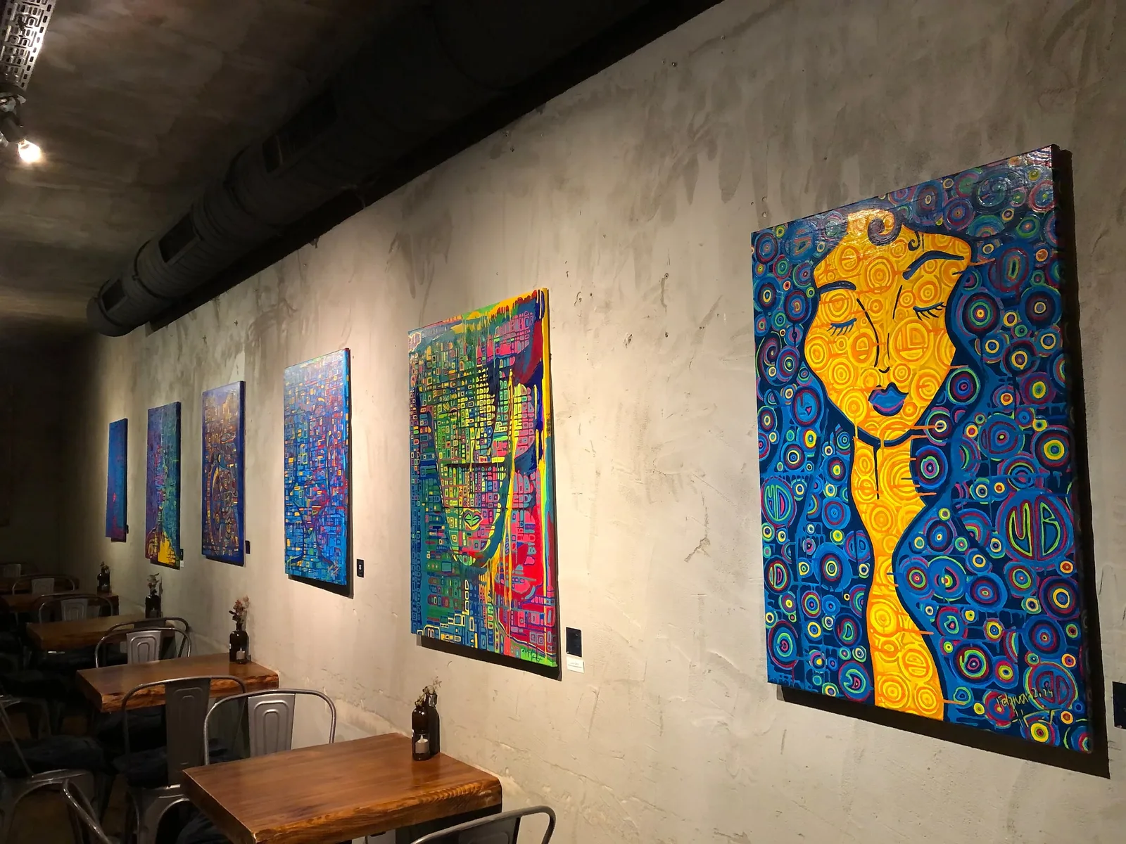 Galerie-Ausstellung mit mehreren abstrakten Kunstwerken an Betonwand, Holztische mit Stühlen