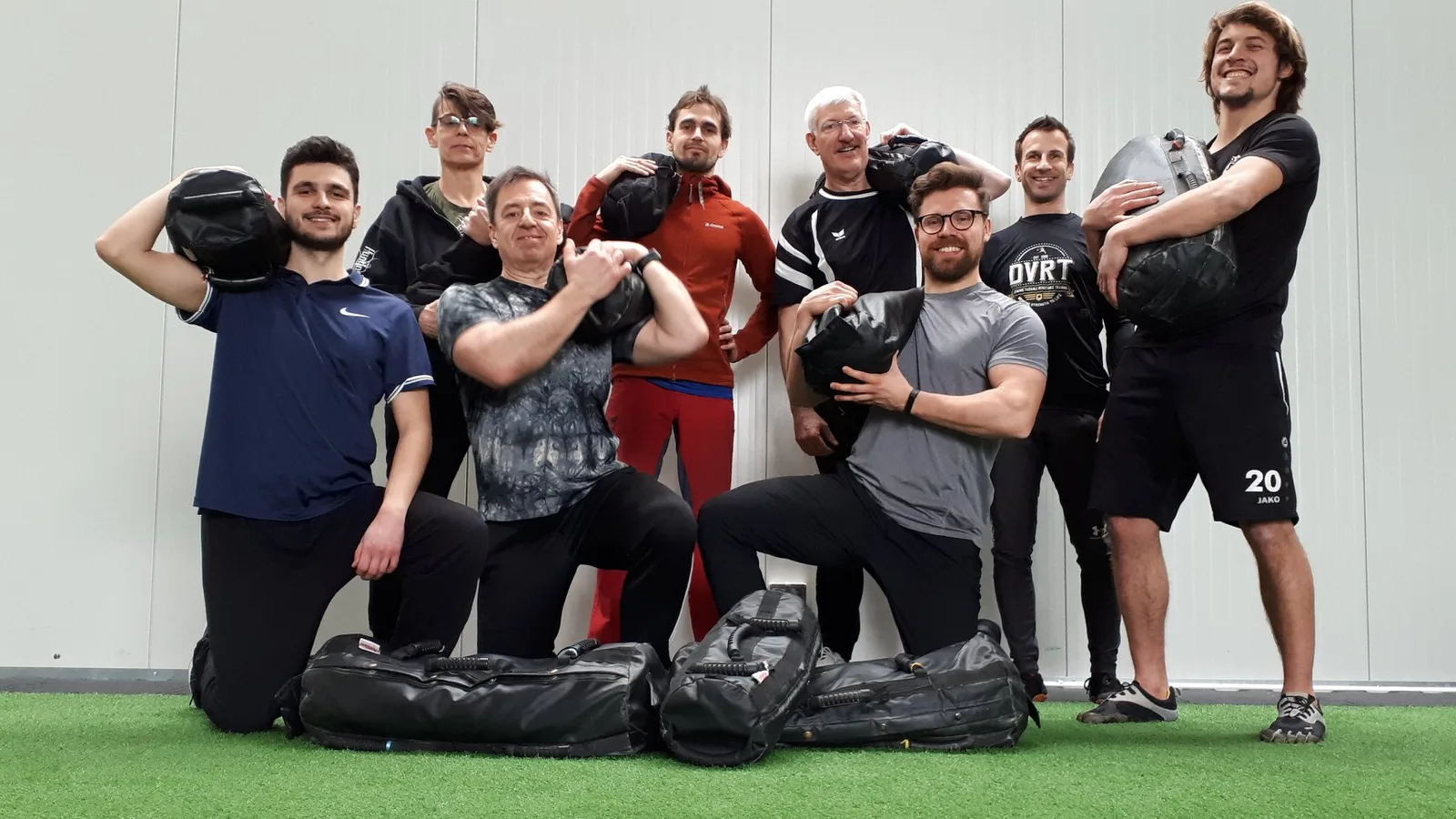 Gruppe von 9 Personen mit Sandsäcken auf Kunstrasenfläche, viele halten Trainingsgeräte