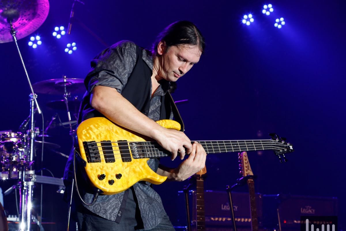 Steffen Knauss konzentriert beim Spielen gelber E-Bass, violettes Bühnenlicht, Schlagzeuger im Hintergrund